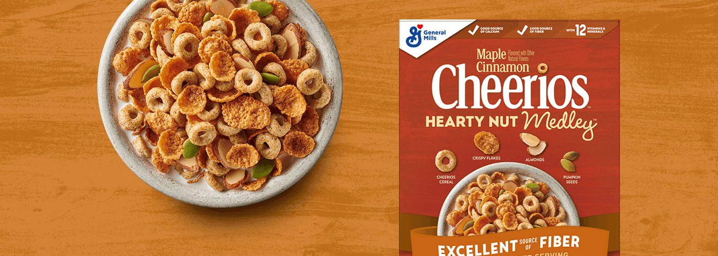 Un tazón de Cheerios Hearty Nut Medley Maple Cinnamon con leche, al lado de la caja de la que provienen. Todo está sobre una tabla de madera.