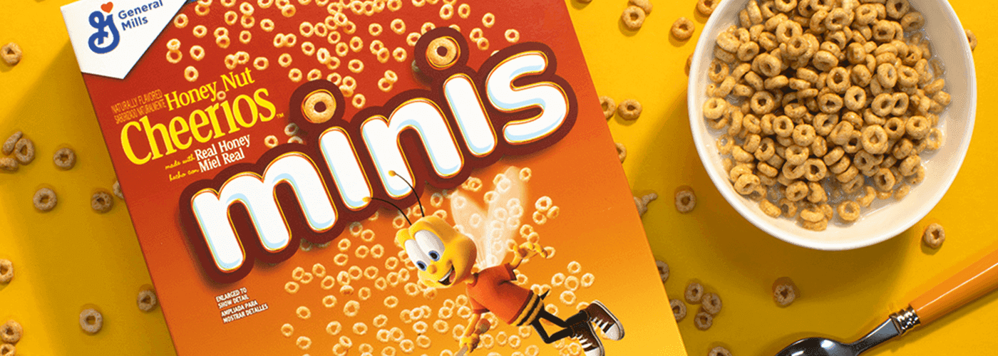Primer plano de una caja de Honey Nut Cheerios Minis junto a un bol de cereales, con una cuchara debajo del bol. Los cereales están esparcidos por la caja y el bol.