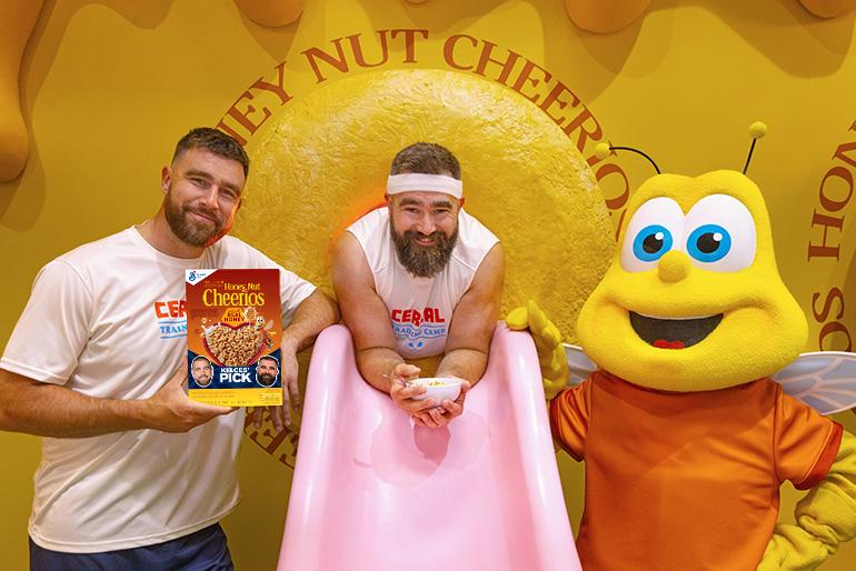 Kelce Bros holding a honey nut cheerios cereal box and a bowl of honey nut cereal and posed with a person in a bee costume in front of a yellow Honey Nut Cheerios backdrop.