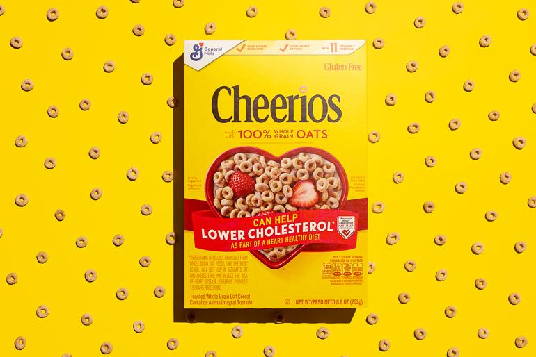 A box of original Cheerios cereal is centered against a bright yellow background patterned with scattered Cheerios pieces. The box features the Cheerios logo, "100% Whole Grain Oats," and a red heart filled with cereal and a strawberry, promoting heart health and cholesterol reduction. It also notes the cereal is gluten-free and displays the General Mills logo in the top left.